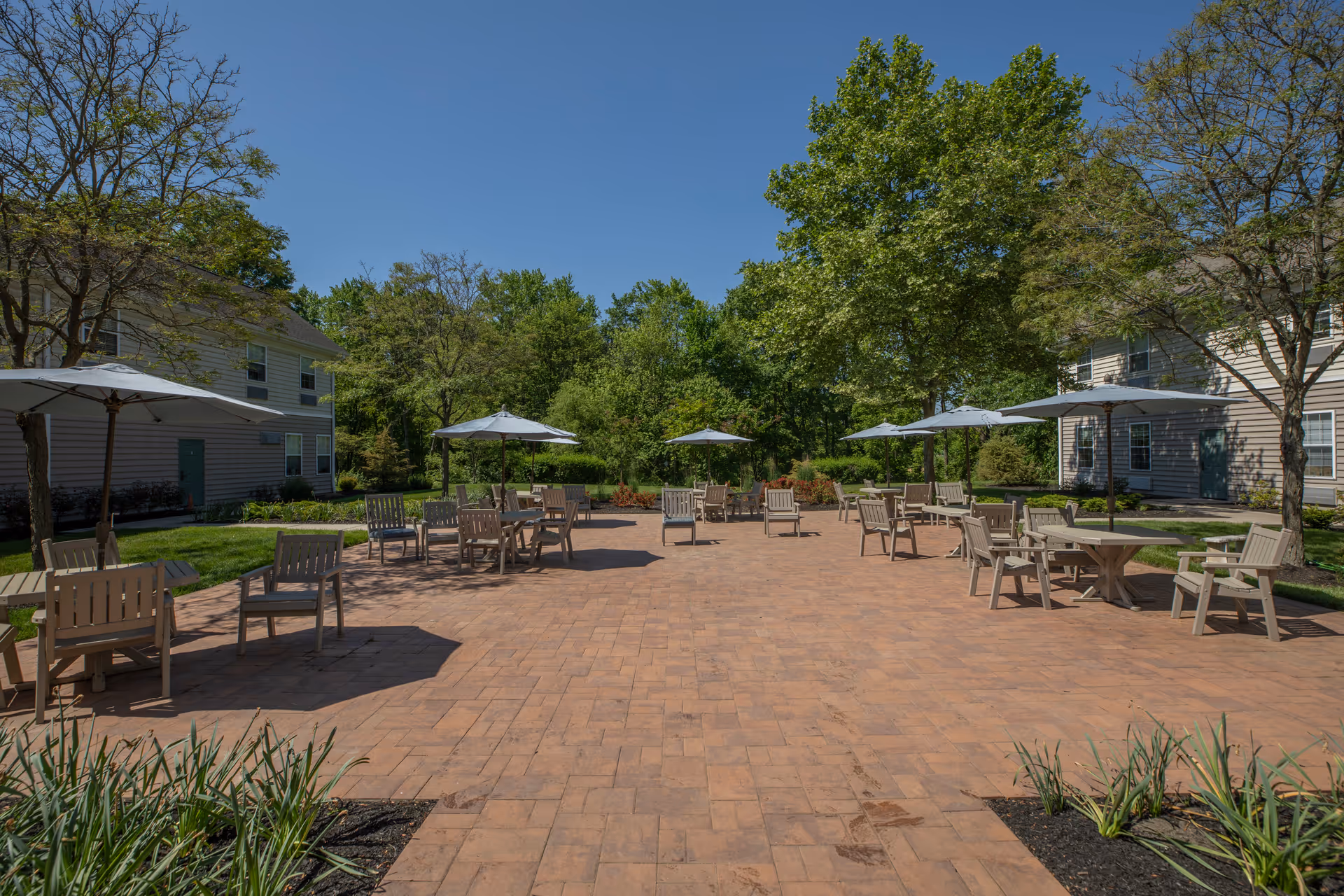 Outdoor patio area with multiple tables and chairs, each shaded by large white umbrellas. The patio is paved with brick tiles and surrounded by green trees and shrubs, with two residential buildings visible on either side under a clear blue sky.