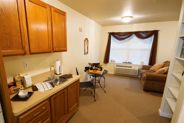 Small studio-style living area with a kitchenette on the left, a round bistro table in the center, and a couch beneath a curtained window.