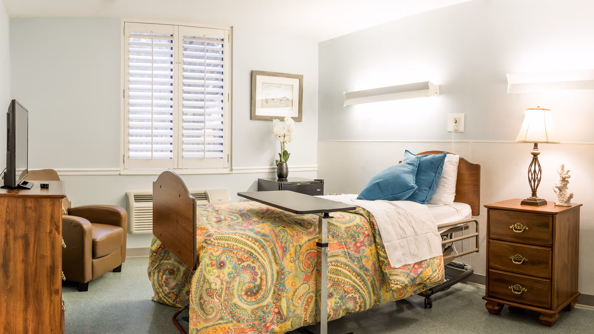 A well-lit senior living facility bedroom with a single bed covered in a colorful paisley-patterned blanket and blue pillows. The room features a wooden nightstand with a lamp and decorative coral, a wooden dresser with a flat-screen TV, a brown armchair, a window with white shutters, and a small table over the bed.