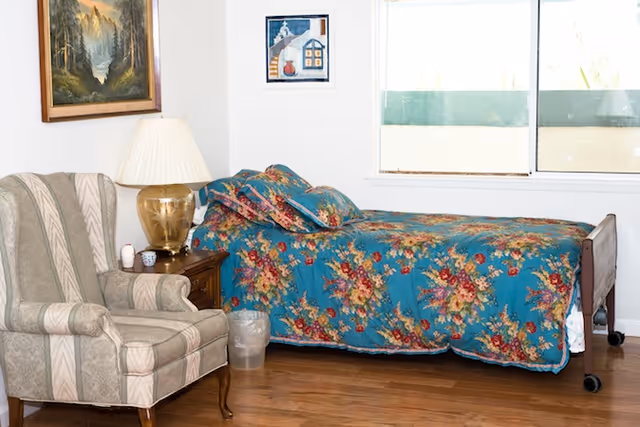 A senior living bedroom with a single bed covered in a blue floral bedspread, a striped upholstered armchair, a wooden nightstand with a gold lamp, and two framed pictures on the white walls. There is a window letting in natural light.