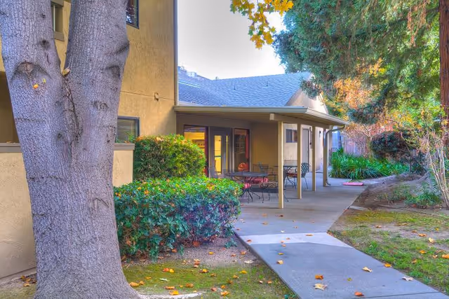 Outdoor patio area of a residential building with a covered seating area featuring tables and chairs. The patio is surrounded by trees, bushes, and a grassy lawn with a concrete walkway leading to the entrance.