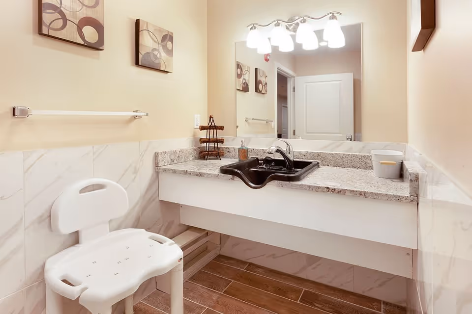 A bathroom with a white plastic shower chair, a granite countertop with a black sink, a large mirror above the sink, and three abstract art pieces on the beige wall. The floor has wood-like tiles and there is a towel rack on the wall.