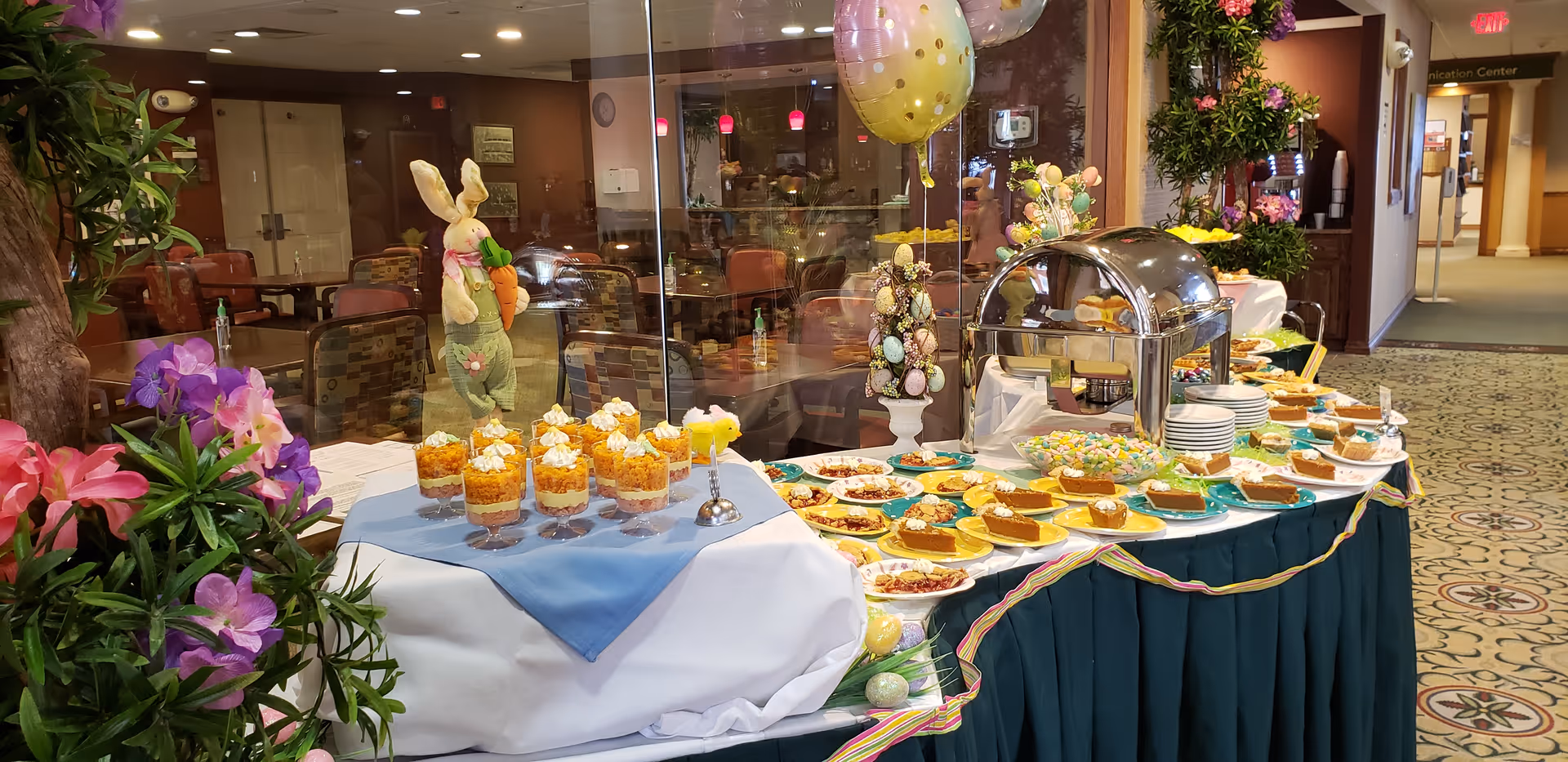 A buffet table set up in an indoor common area with various desserts including pumpkin pie slices, parfaits topped with whipped cream, and other sweet treats. The table is decorated with Easter-themed items such as a bunny figure holding a carrot, pastel-colored eggs, and balloons. There are flowers and greenery around the table, and chairs and tables are visible in the background.