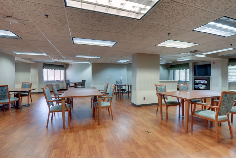 A spacious dining room with several wooden tables and chairs arranged neatly on a wood floor. The room has a drop ceiling with fluorescent lighting, windows with blinds and valances, and a TV on a stand in the corner.