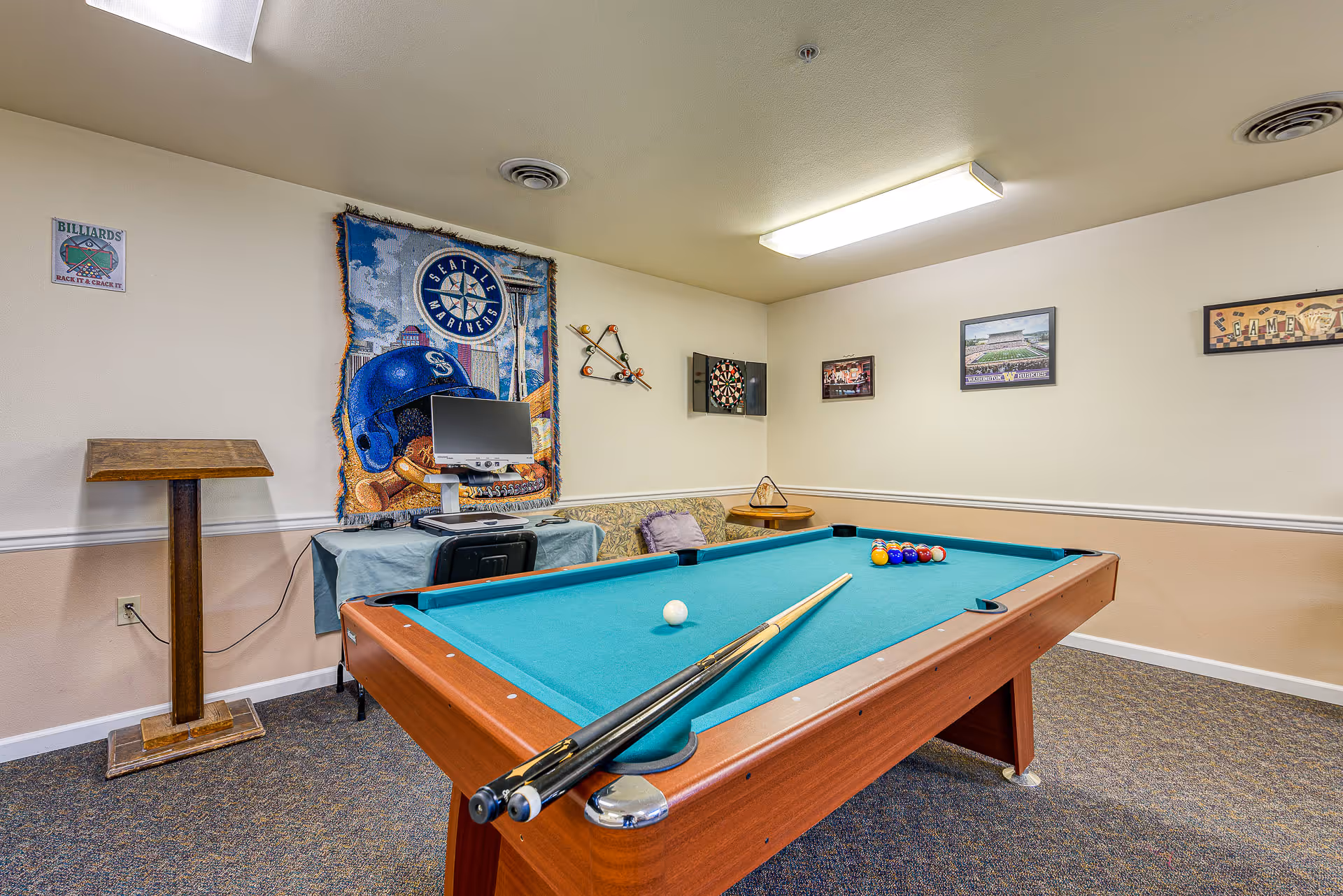 A recreational room with a pool table in the center, two pool cues resting on the table, and a set of billiard balls arranged for a game. The walls are decorated with sports-themed pictures and a Seattle Mariners tapestry. There is a dartboard mounted on the wall and a small table with a computer monitor and chair in the corner. The room has beige walls, carpeted floor, and fluorescent ceiling lights.