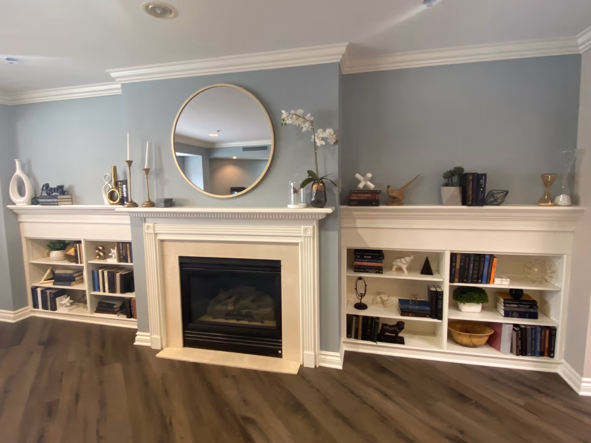 A cozy living room area featuring a white fireplace with a beige surround, flanked by built-in white bookshelves filled with books and decorative items. Above the fireplace is a round mirror with a gold frame, and the walls are painted a soft blue-gray. The floor is a dark wood laminate.