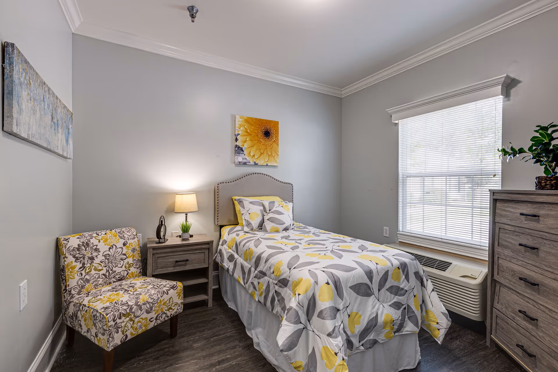 A small, neatly arranged bedroom with a single bed covered in a floral-patterned bedspread in shades of yellow, gray, and white. Next to the bed is a wooden nightstand with a lamp and a small plant. A floral-patterned chair is placed against the wall on the left side. There is a window with blinds on the right side, and a wooden dresser with a plant on top is visible. The walls are painted light gray and decorated with two pieces of wall art.