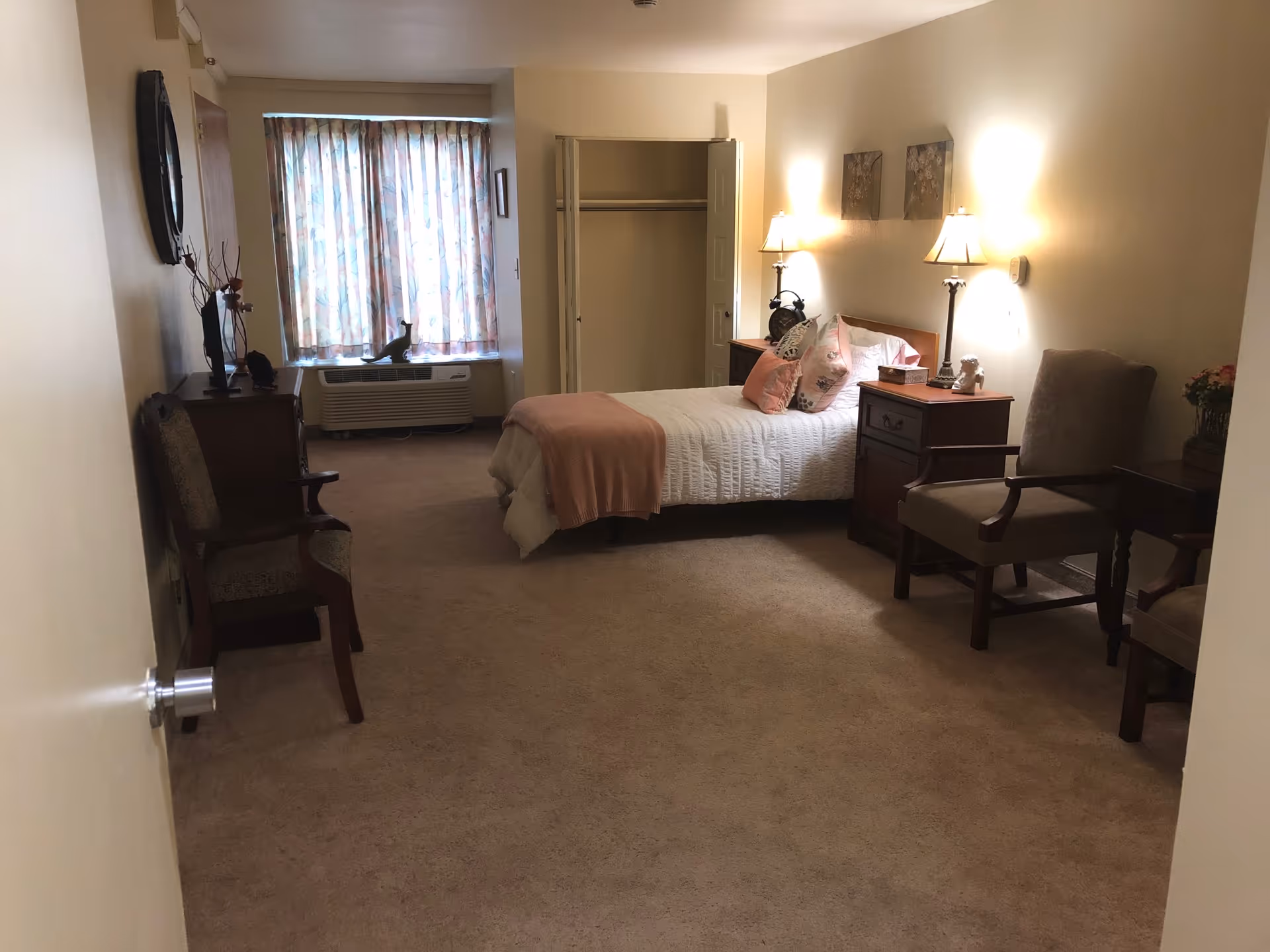 A cozy bedroom in a senior living facility with a single bed covered in white bedding and a pink throw blanket. The room has two bedside tables with lamps, two armchairs, a small table, a dresser with a TV, and a window with floral curtains. The closet door is open, and the walls are decorated with two framed pictures.