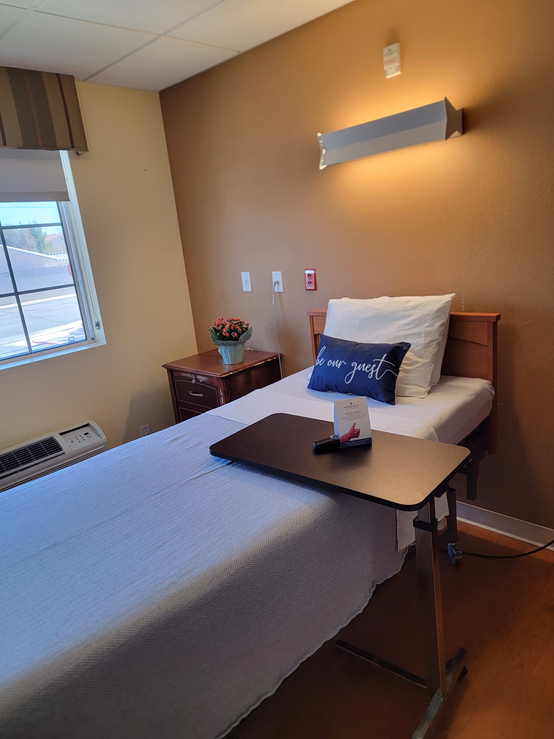 Single patient bed with overbed table, pillows, and window in a small beige bedroom.
