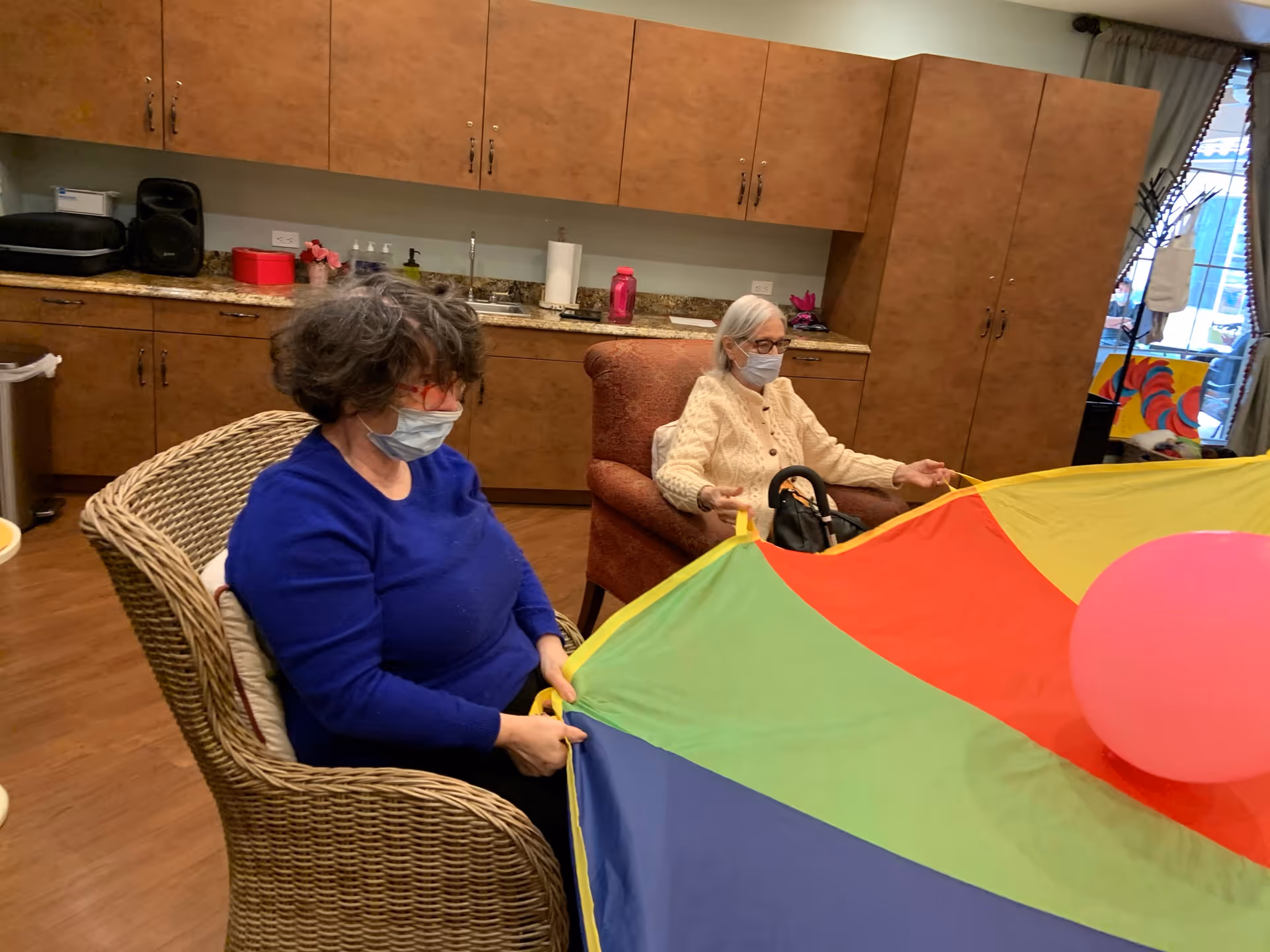 Two elderly women wearing face masks are seated in a room with wooden cabinets and a countertop. They are holding the edges of a colorful parachute with a pink balloon on top. One woman is in a wicker chair wearing a blue sweater, and the other is in a cushioned armchair wearing a cream-colored sweater.