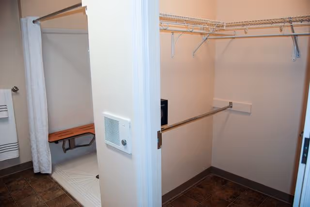 View of a bathroom and adjacent walk-in closet. The bathroom features a shower area with a white curtain and a wooden fold-down bench. The closet has wire shelving and a hanging rod. The floor is tiled in both areas.