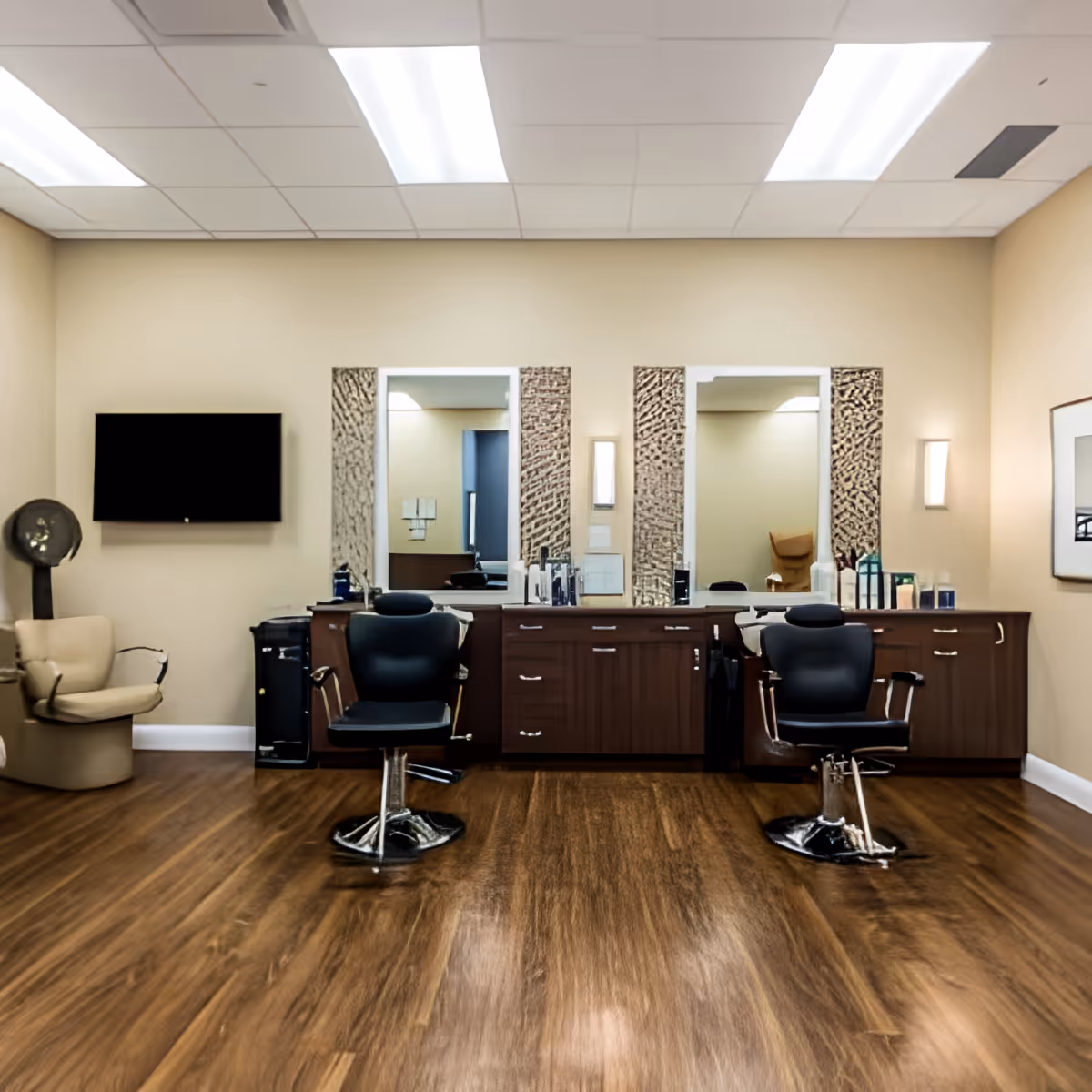 Interior view of a salon area in a senior living facility with two black salon chairs in front of a wooden counter with mirrors. The room has beige walls, wood flooring, a beige armchair, a wall-mounted TV, and various hair care products on the counter.