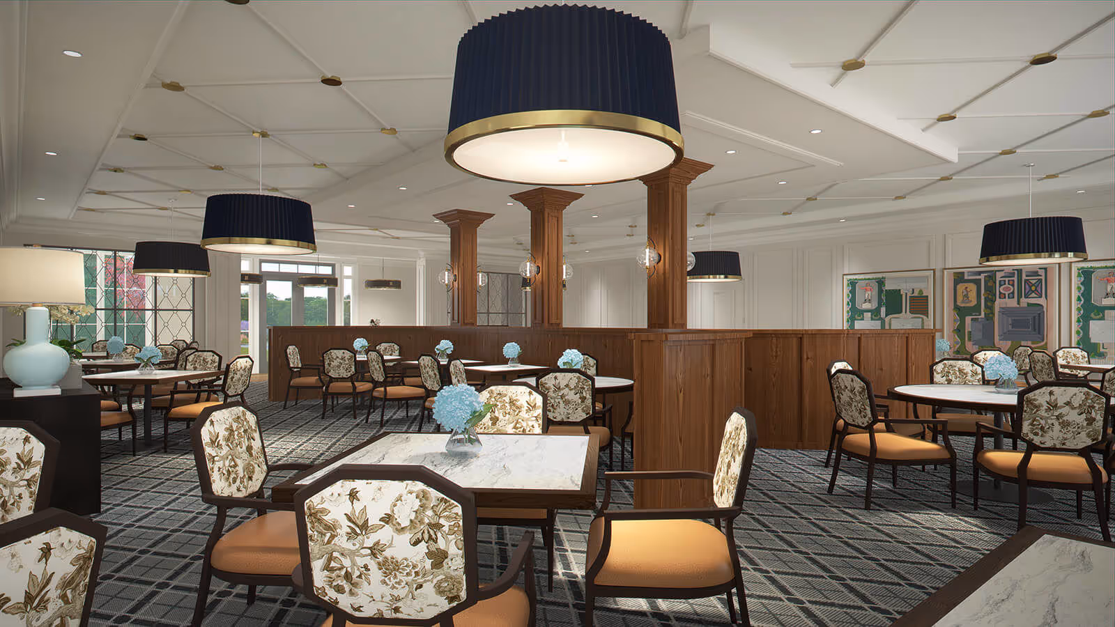 A spacious dining room with multiple round and square tables, each surrounded by floral-patterned chairs with orange cushions. The room features a patterned carpet, wooden pillars, and large black pendant lights hanging from a white ceiling. Small blue flower arrangements are placed on each table, and there are decorative lamps and framed artwork on the walls.