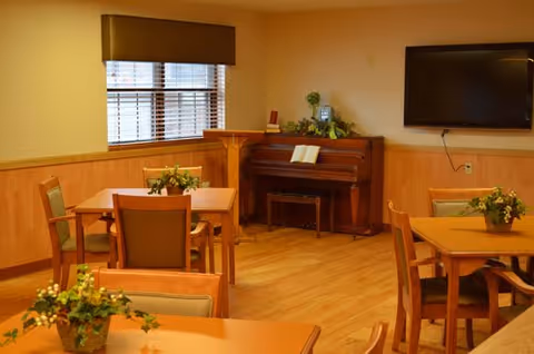 A cozy interior room with wooden floors and walls, featuring several tables and chairs arranged for dining or socializing. A piano with an open book on its stand is placed against the wall, and a flat-screen TV is mounted on the wall nearby. Potted plants decorate the tables and the piano area.