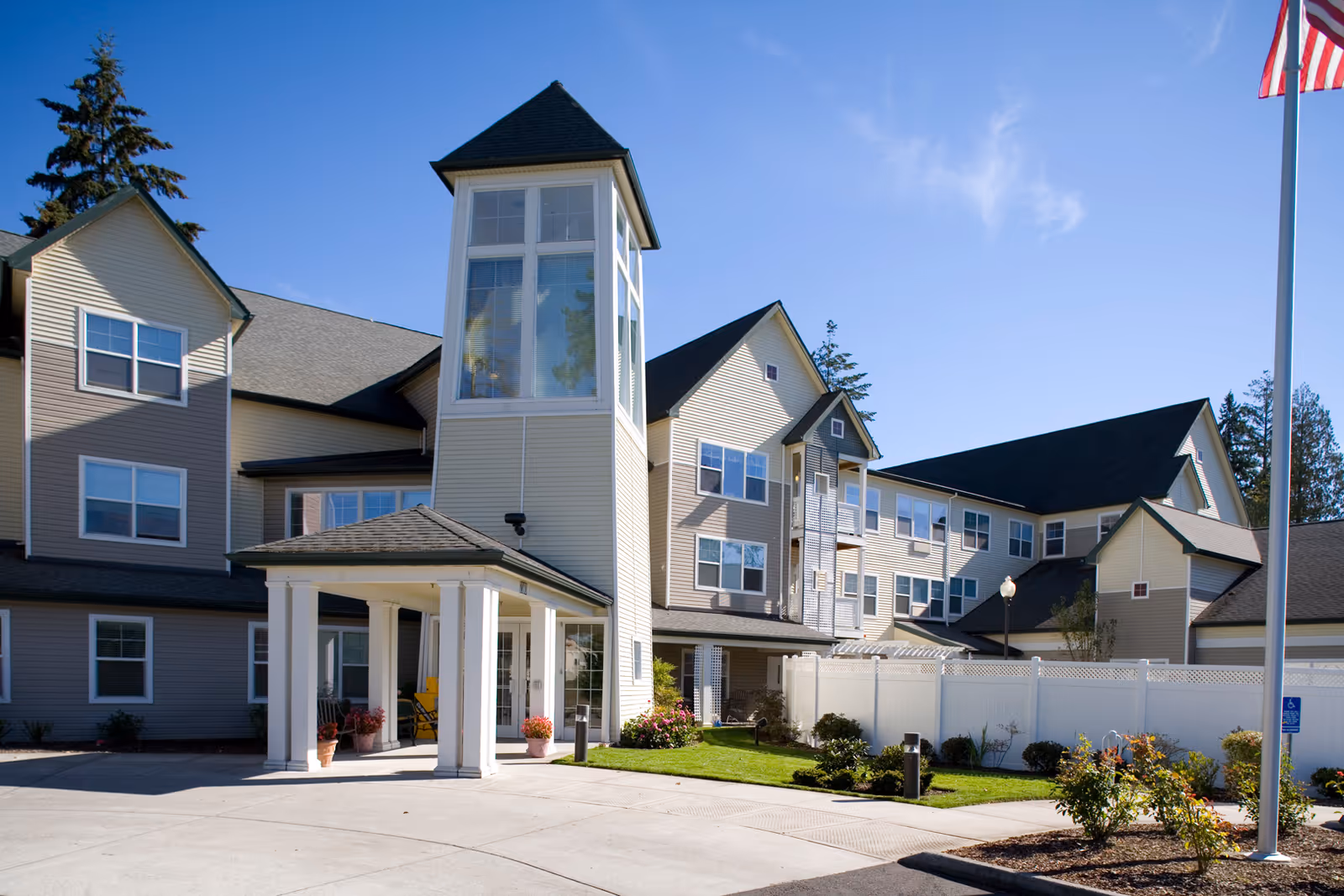 Exterior view of Avamere at Hillsboro senior living facility showing a multi-story building with beige siding, a covered entrance supported by white columns, landscaped greenery, and a flagpole with an American flag against a clear blue sky.