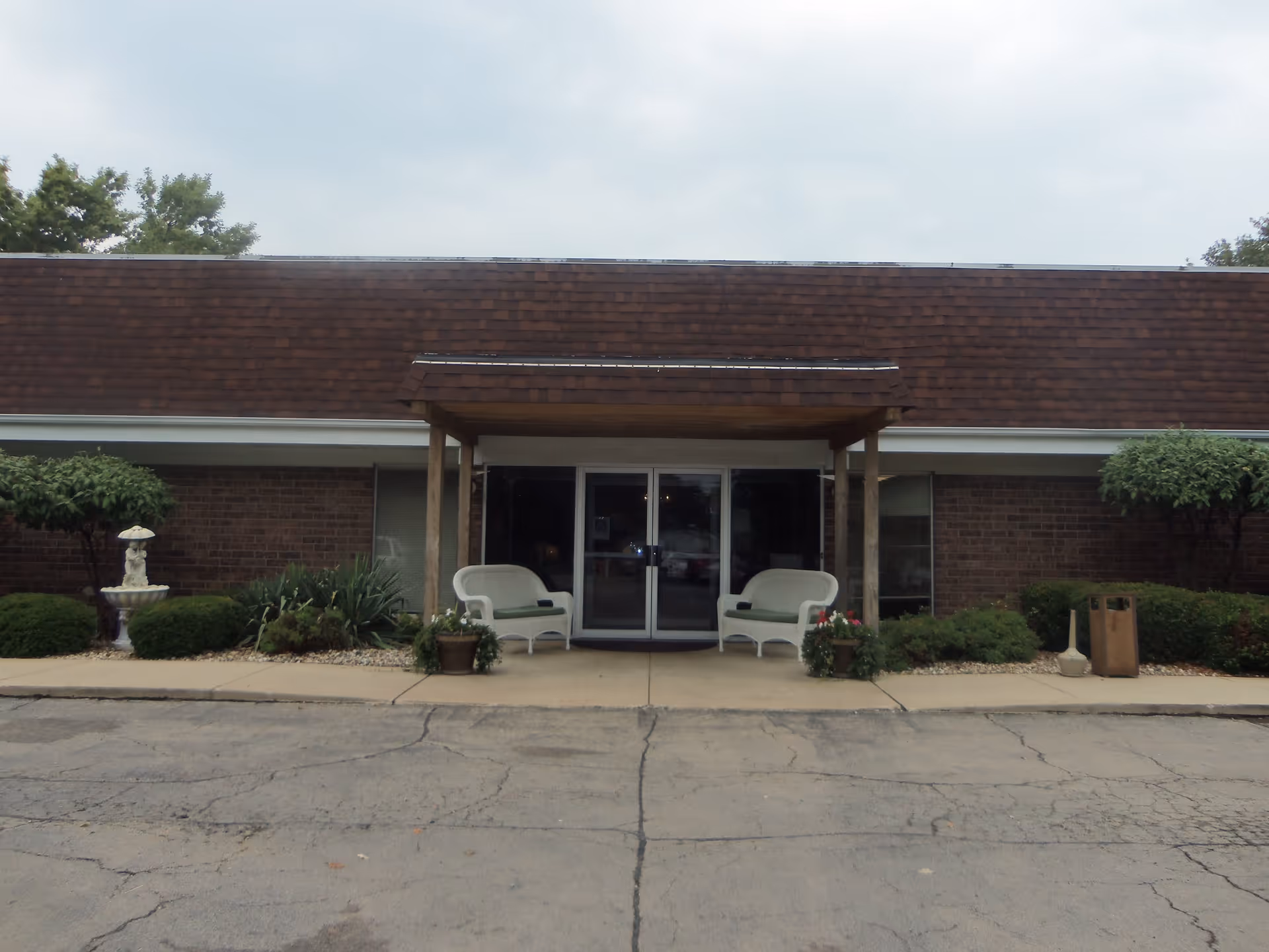 Front entrance of a single-story brick building with a brown shingled roof. The entrance has a small covered porch with two white benches and potted plants on either side. There are bushes and small trees along the front of the building, and a decorative fountain is visible on the left side.