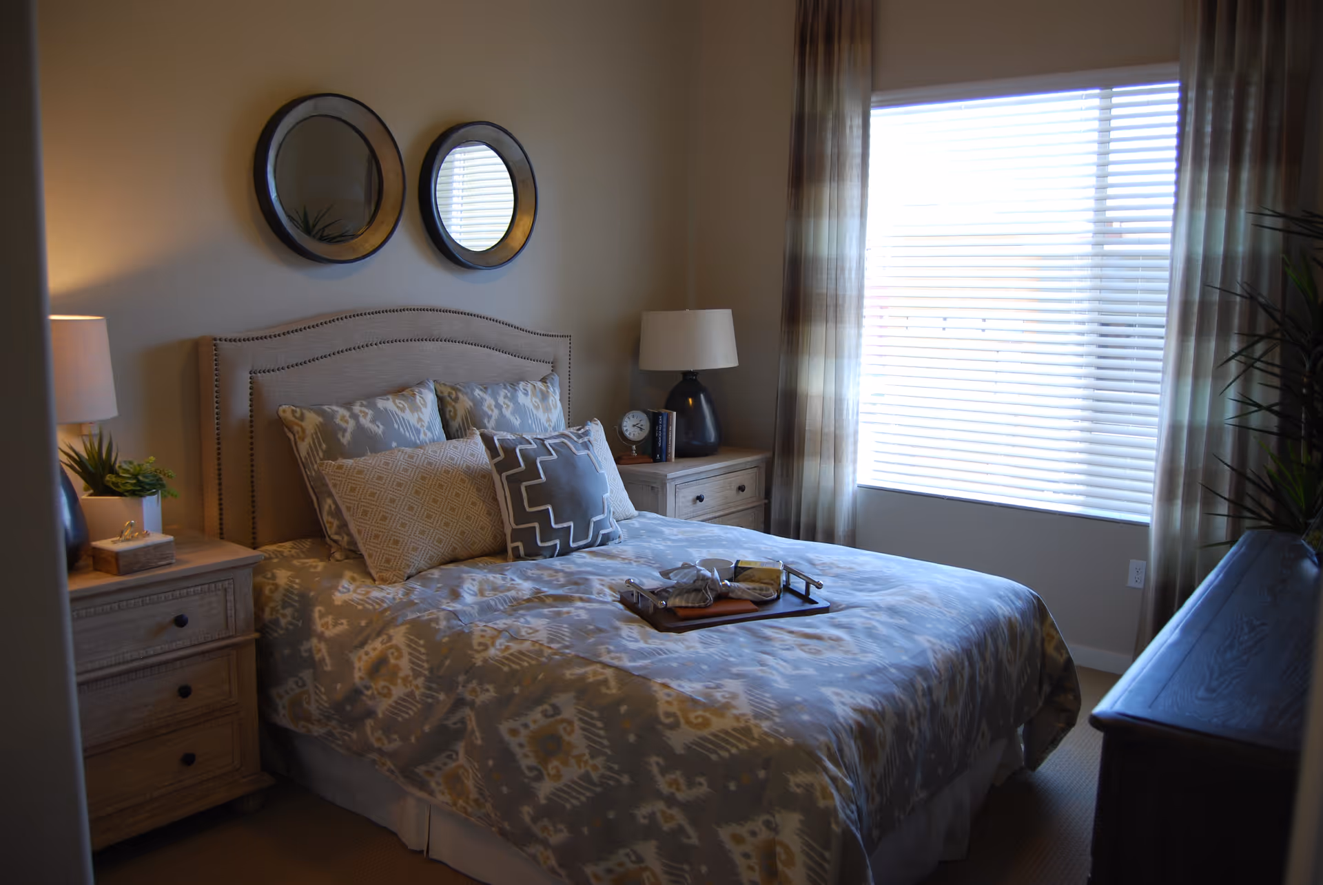 A sunlit furnished bedroom with a made bed, patterned bedding, matching nightstands and lamps, and two round mirrors above the headboard.