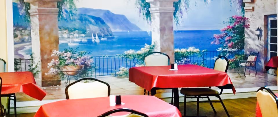 Dining area with tables covered in red tablecloths and chairs around them. The background features a large mural depicting a scenic coastal view with blue water, sailboats, mountains, and flowering plants on a terrace with stone columns.