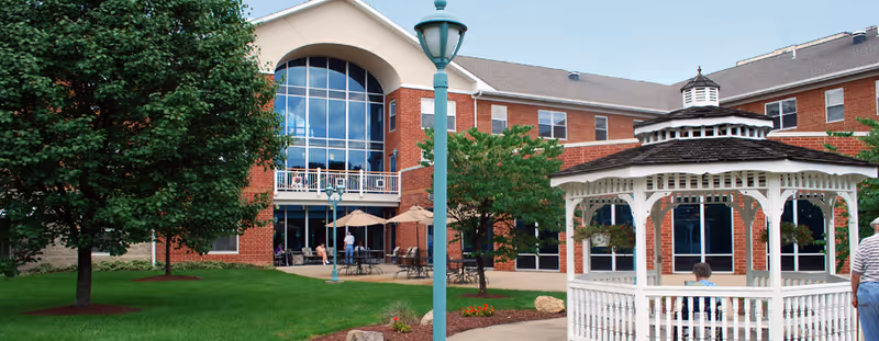 Outdoor view of Paramount Senior Living South Hills showing a red brick building with large windows and a balcony. In the foreground, there is a white gazebo with a person sitting inside, green grass, trees, and outdoor seating with umbrellas.