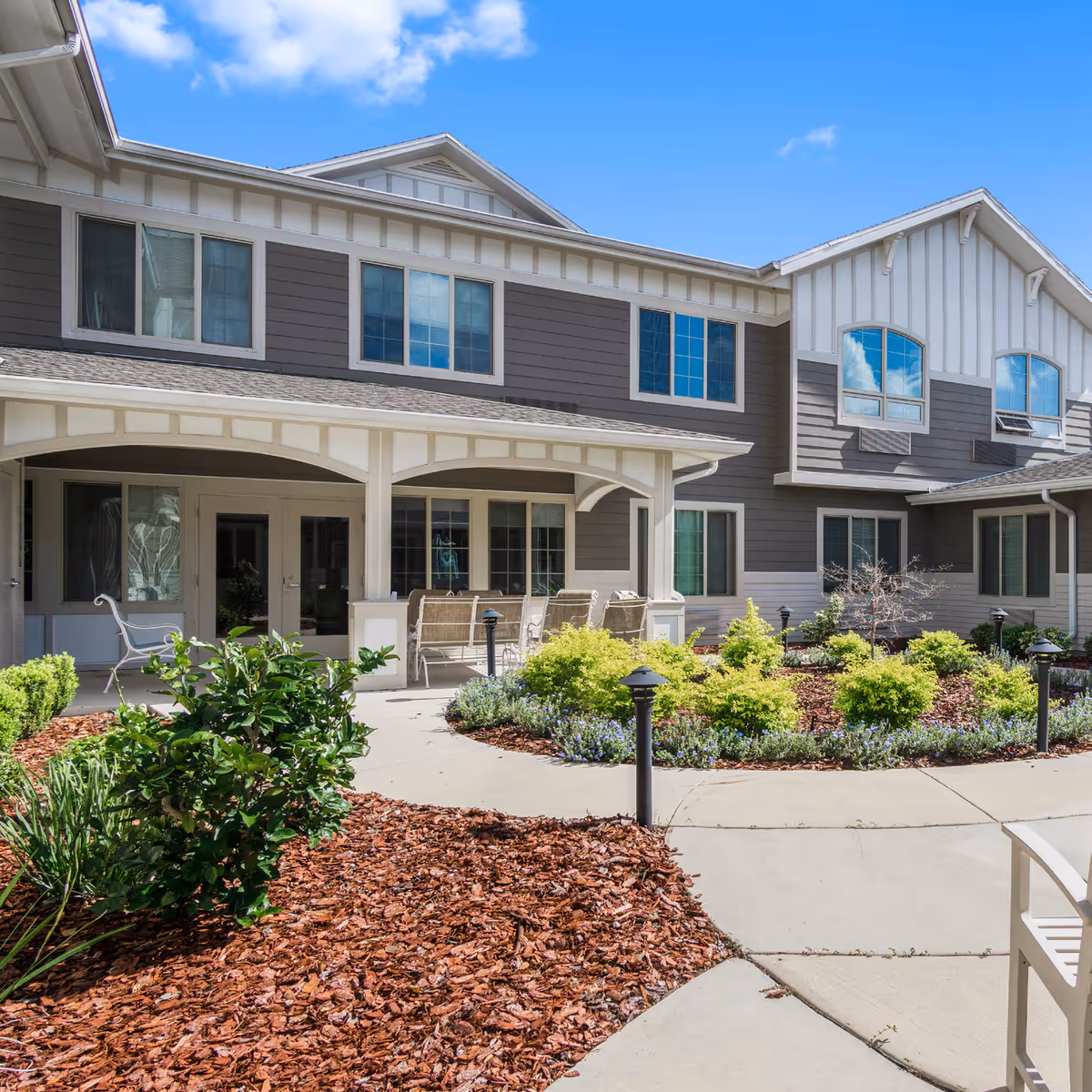 Exterior view of The Willows senior living facility showing a two-story building with gray and white siding, multiple windows, and a covered patio area with outdoor seating. The foreground features a landscaped garden with shrubs, mulch, and a curved concrete walkway under a bright blue sky.