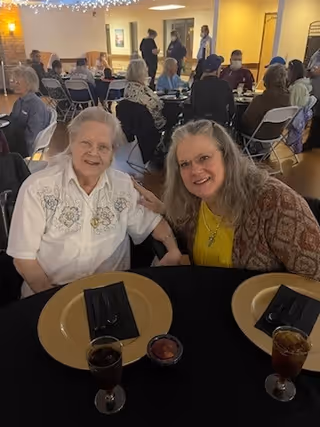 Two elderly women sitting at a round table set with gold chargers, black napkins, and glasses of iced tea in a dining area with other people seated at tables in the background.