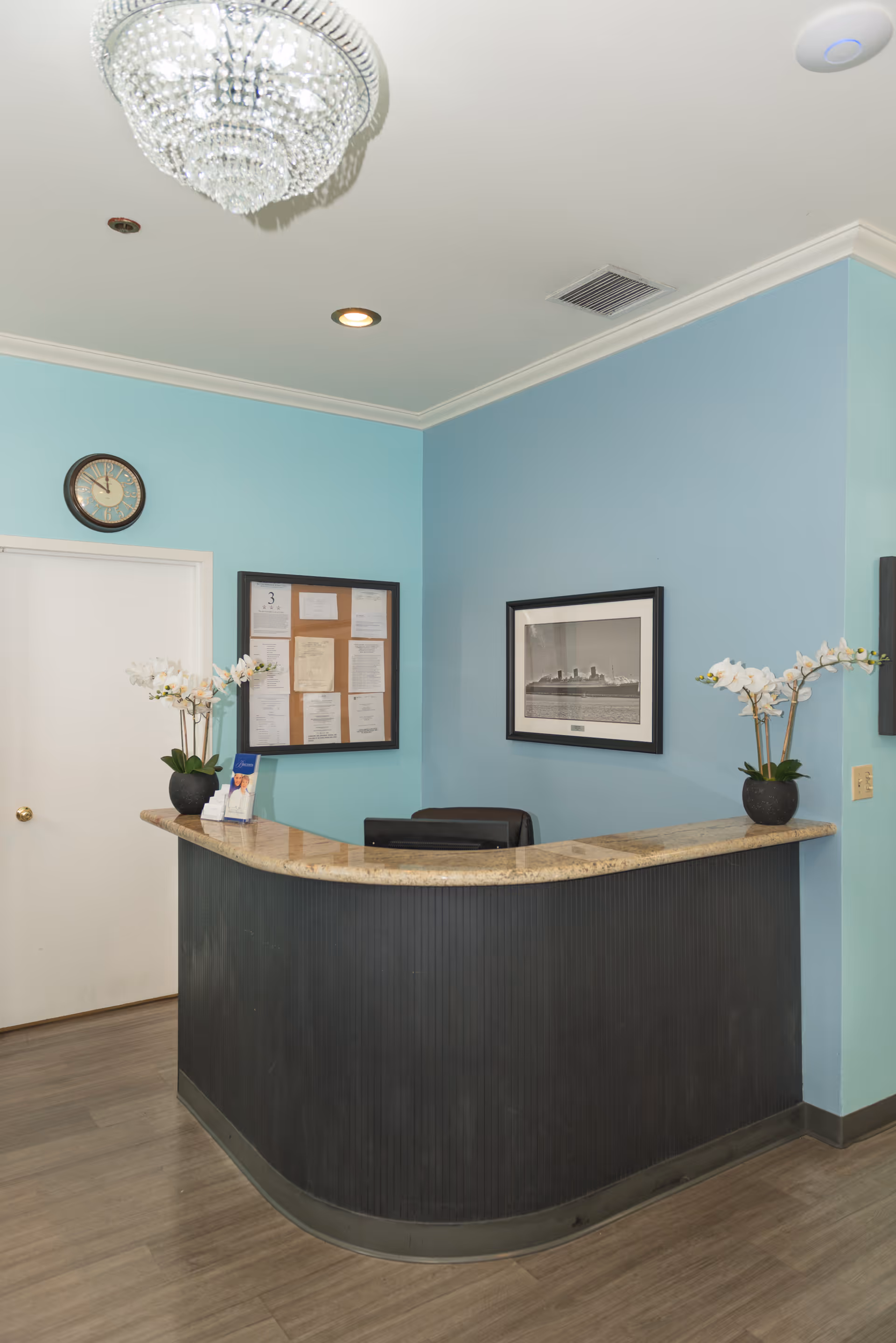 Reception area with a curved black front desk topped with beige marble. Two potted white orchid plants are placed on the desk. The walls are painted light blue and decorated with a framed black and white photograph and a bulletin board with papers. A round wall clock is above a closed white door. The ceiling has a crystal chandelier and recessed lighting.