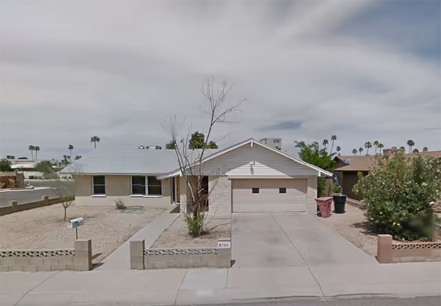Single-story residential building with a light-colored exterior, a two-car garage, a driveway, and a small front yard with desert landscaping including gravel and a few sparse trees and bushes. The sky is cloudy and there are palm trees visible in the background.
