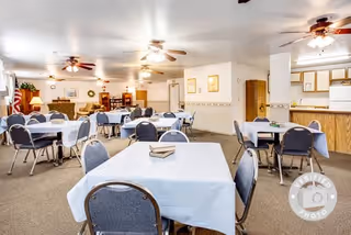 A spacious dining room with multiple tables covered in white tablecloths, each surrounded by blue cushioned chairs. The room is well-lit with ceiling fans and lights, and there is a kitchen area visible in the background with wooden cabinets and white appliances. The walls are decorated with framed pictures and a clock.