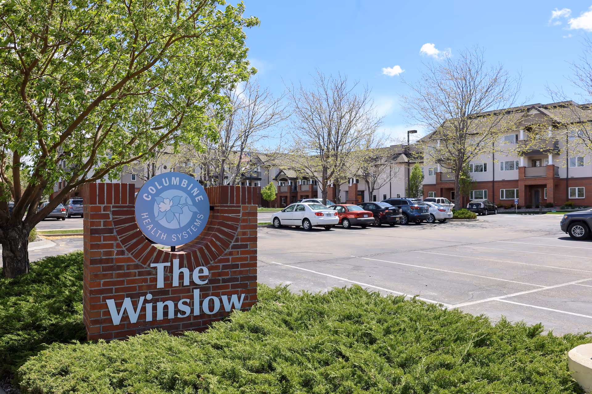 Exterior view of The Winslow Independent Living facility with a brick sign in the foreground that reads 'Columbine Health Systems The Winslow'. The sign is surrounded by green shrubs and a tree with green leaves. In the background, there is a parking lot with several cars and a multi-story residential building with balconies and windows under a clear blue sky.
