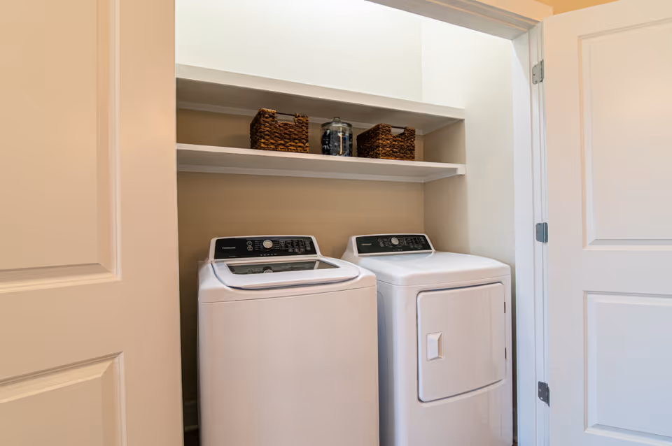 Laundry area with a white top-loading washing machine and a white front-loading dryer side by side, with two white shelves above holding two woven baskets and a glass jar.