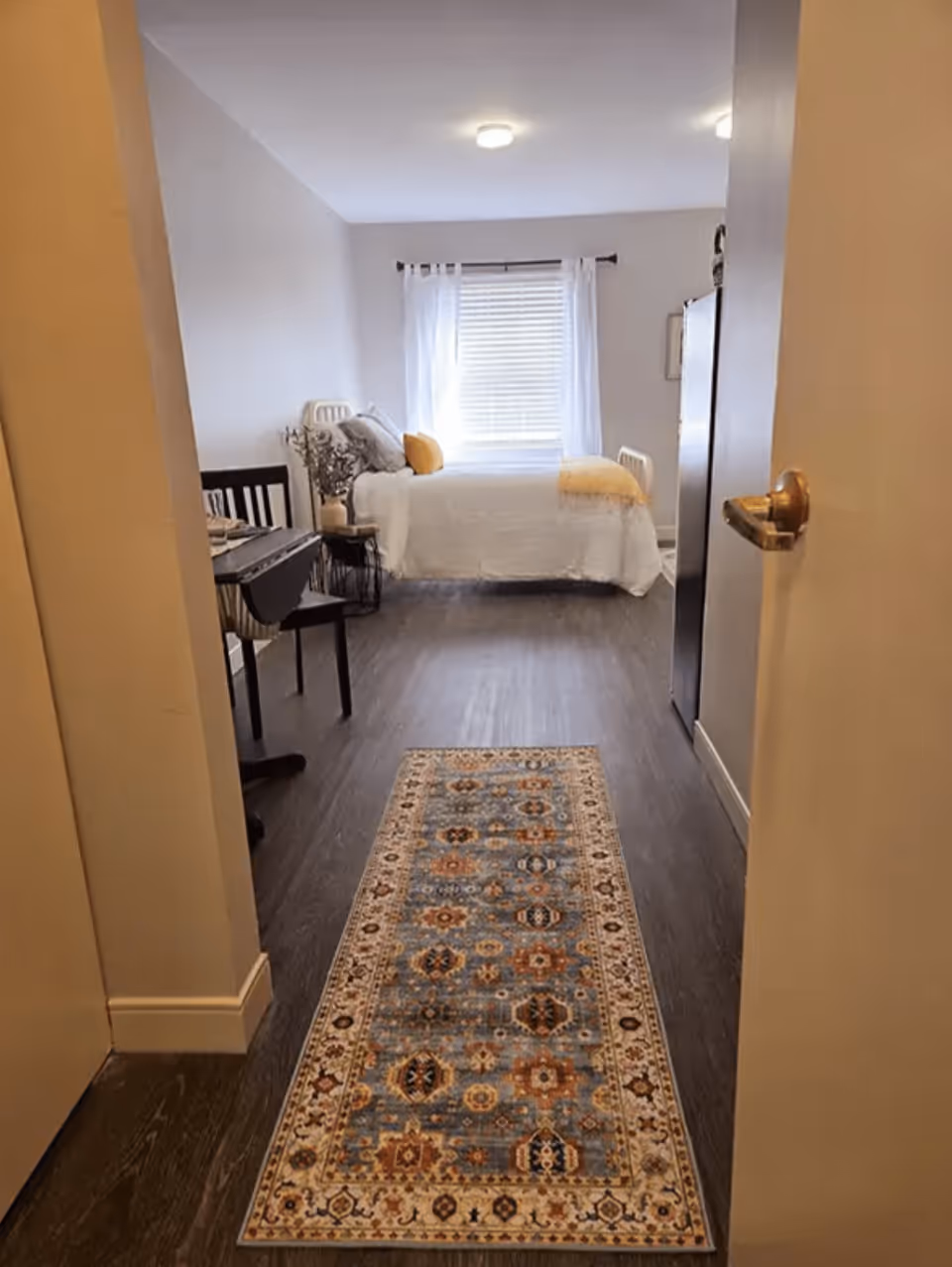 View through a doorway into a bedroom with a bed against the far wall, decorated with white bedding and yellow accent pillows. A window with white curtains is behind the bed. A small table and chair are on the left side, and a patterned rug runs along the dark wood floor leading into the room.
