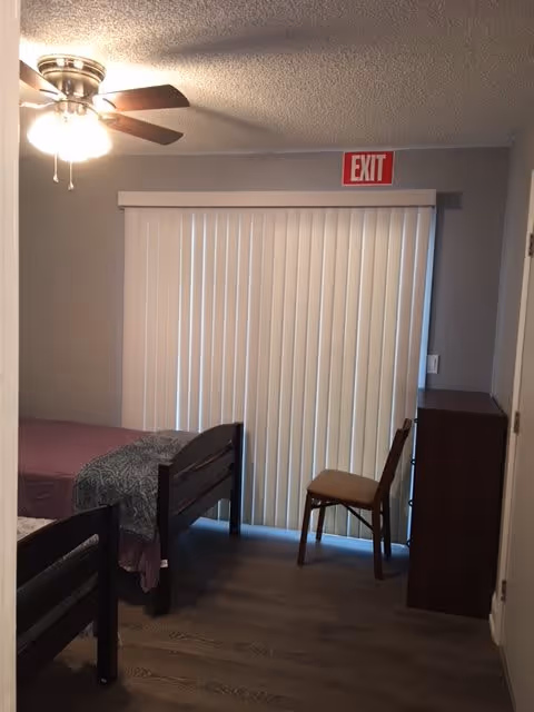 A small bedroom with two single beds, one with a purple blanket and the other with a gray patterned blanket. There is a wooden chair and a dark wooden dresser near a sliding glass door covered with vertical blinds. A ceiling fan with lights is on the ceiling, and an exit sign is above the sliding door.