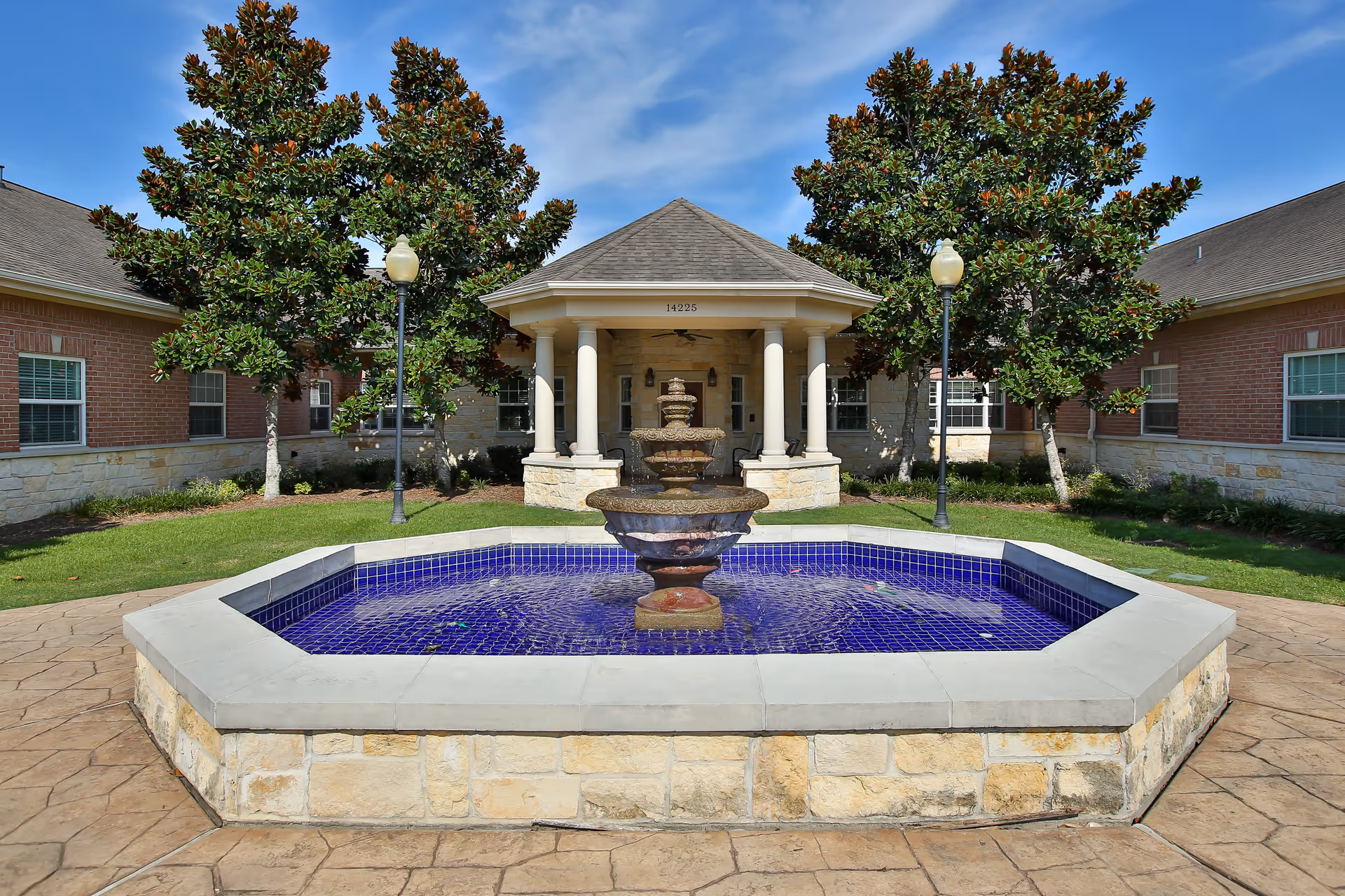 Outdoor courtyard area of a senior living facility featuring a central stone fountain with blue tile lining, surrounded by a paved walkway, green grass, and trees. The building has brick and stone walls with multiple windows and a covered entrance with columns.