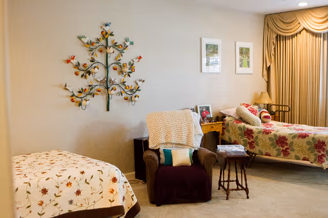 A cozy bedroom with two beds covered in floral bedspreads, a comfortable armchair with a knitted throw and pillows, a small wooden side table, framed pictures on the wall, and a decorative metal tree wall art. The room has beige walls and carpet, with a window covered by beige curtains.