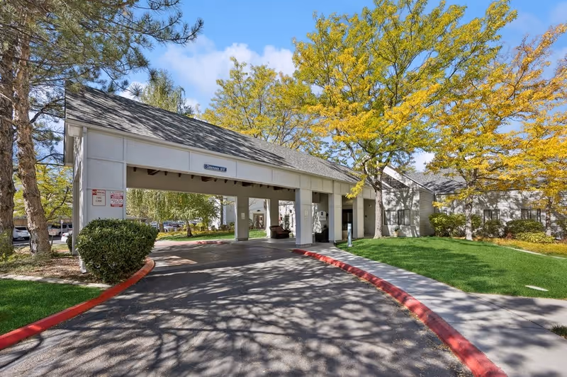 Entrance driveway to a senior living facility with a covered drop-off area, surrounded by green grass, trees with yellow leaves, and a clear blue sky.