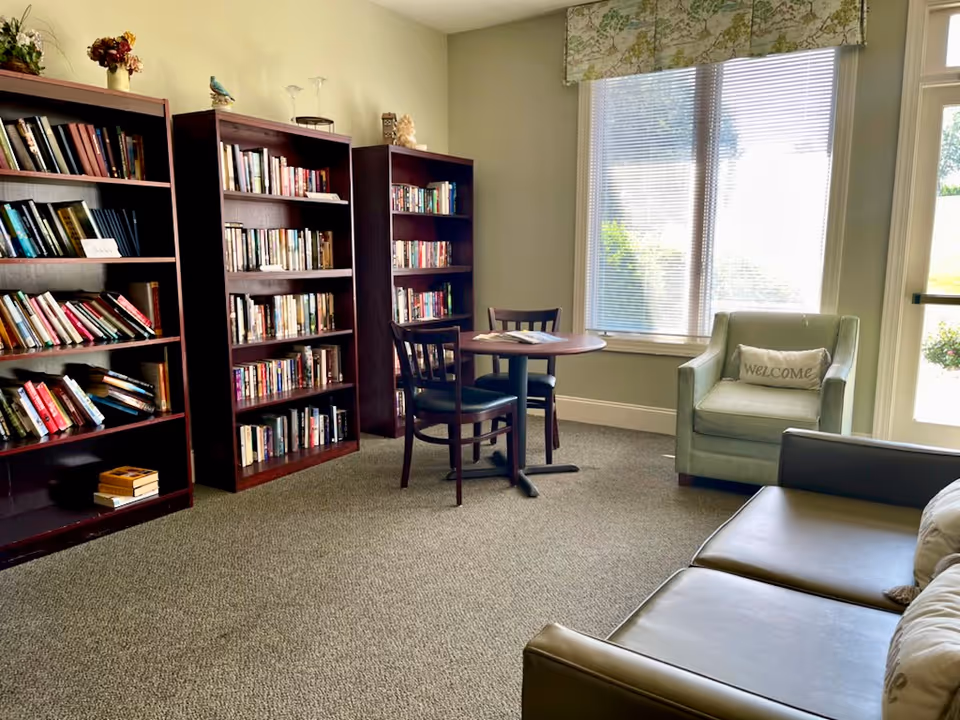 Sunny common room with bookshelves, a round table and chairs, an armchair with a "WELCOME" pillow, and a couch.