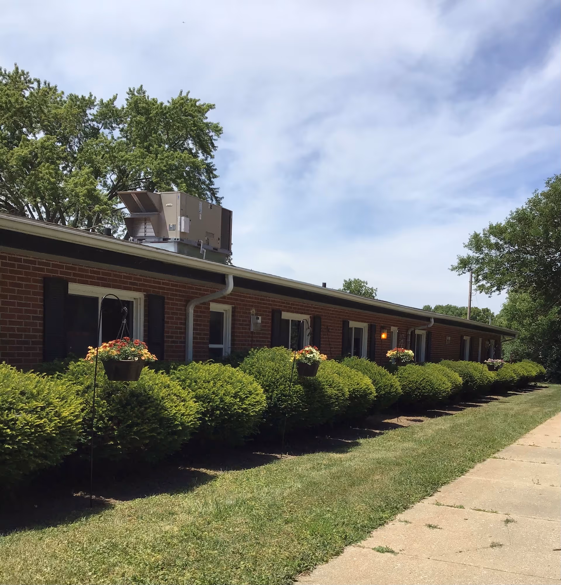 Exterior view of a single-story brick building with several windows, black shutters, and a row of neatly trimmed bushes in front. Hanging flower pots with colorful flowers are placed along the bushes. The sky is partly cloudy with some trees visible in the background.
