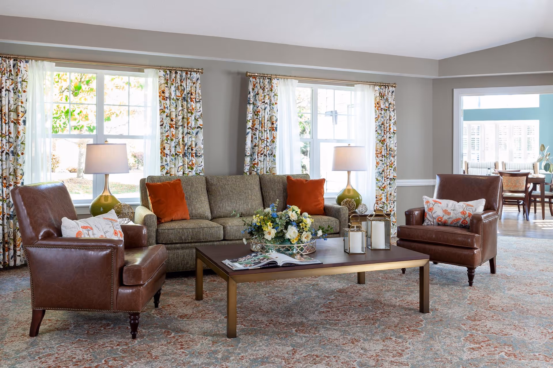 A cozy living room with a gray sofa adorned with orange pillows, two brown leather armchairs with floral cushions, a wooden coffee table with a floral centerpiece and lanterns, and large windows with floral curtains letting in natural light. In the background, there is a dining area with chairs and a table.