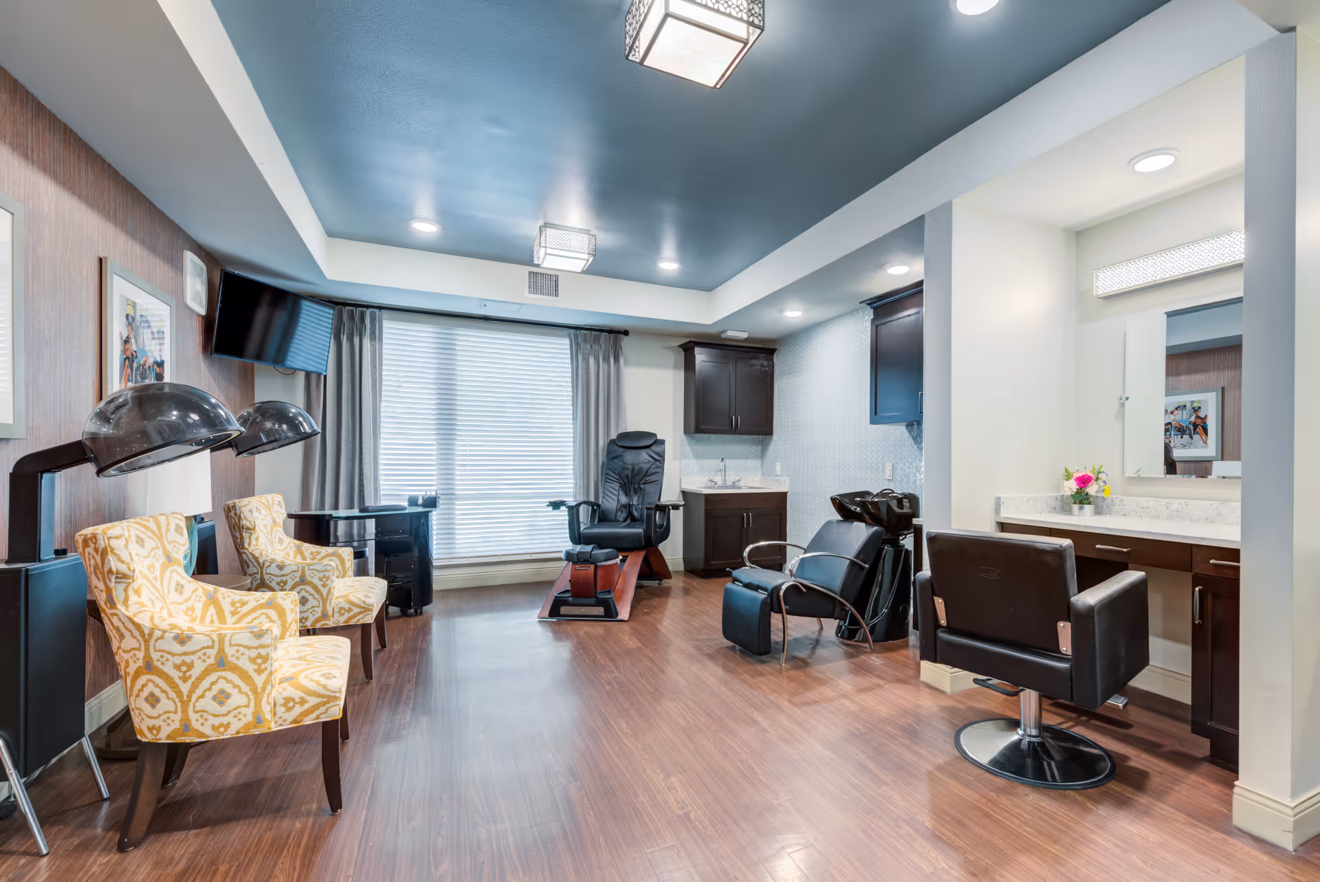 Interior view of a salon area in a senior living facility with two yellow patterned chairs under hair dryers on the left, a black pedicure chair in the center, a black shampoo station with a chair on the right, and a vanity with a mirror and flower vase. The room has wood flooring, a blue ceiling, and large window with blinds and curtains.