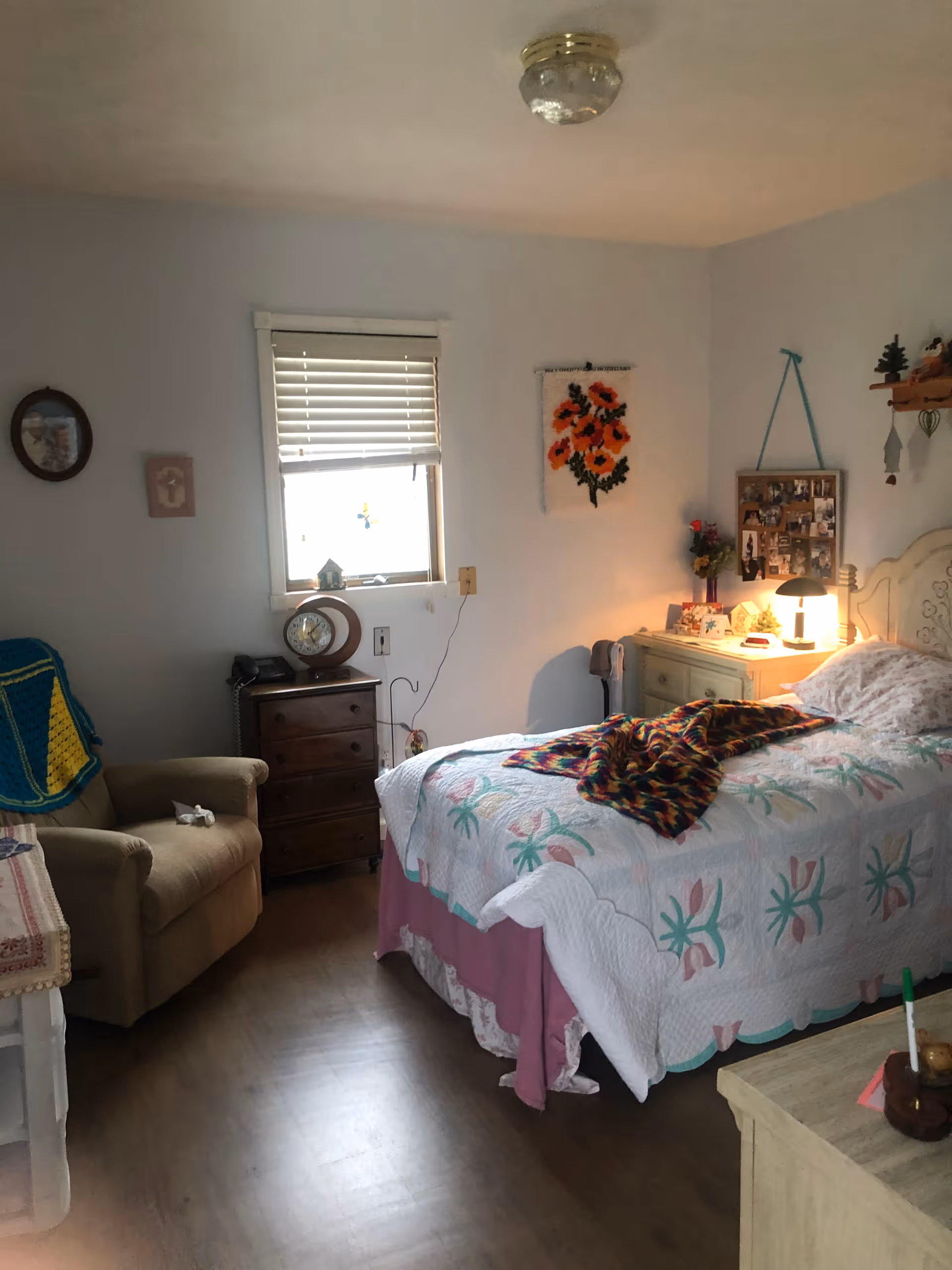 A cozy bedroom in an assisted living facility featuring a single bed with a floral quilt and a colorful knitted blanket. Next to the bed is a nightstand with a lamp, flowers, and personal items. There is a wooden dresser with a clock and phone under a window with blinds. An armchair with a crocheted blanket is positioned near the dresser. The walls have framed pictures and a hanging floral tapestry. The room has wooden flooring and soft lighting.