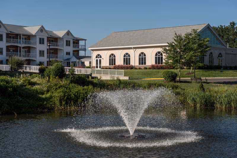 A scenic view of a senior living facility with a large pond featuring a water fountain in the foreground. Behind the pond, there are two buildings, one with multiple balconies and the other with large arched windows. The area is surrounded by greenery and trees under a clear blue sky.