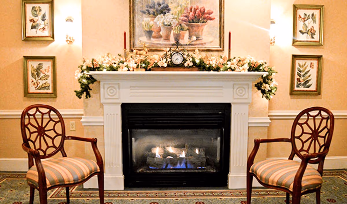 A cozy interior scene featuring a white fireplace with a fire burning inside. On the mantel above the fireplace, there is a floral garland, a small clock, two red candles, and a framed painting of flowers. Two wooden chairs with striped cushions are positioned on either side of the fireplace. The walls are decorated with framed botanical prints and soft lighting fixtures.