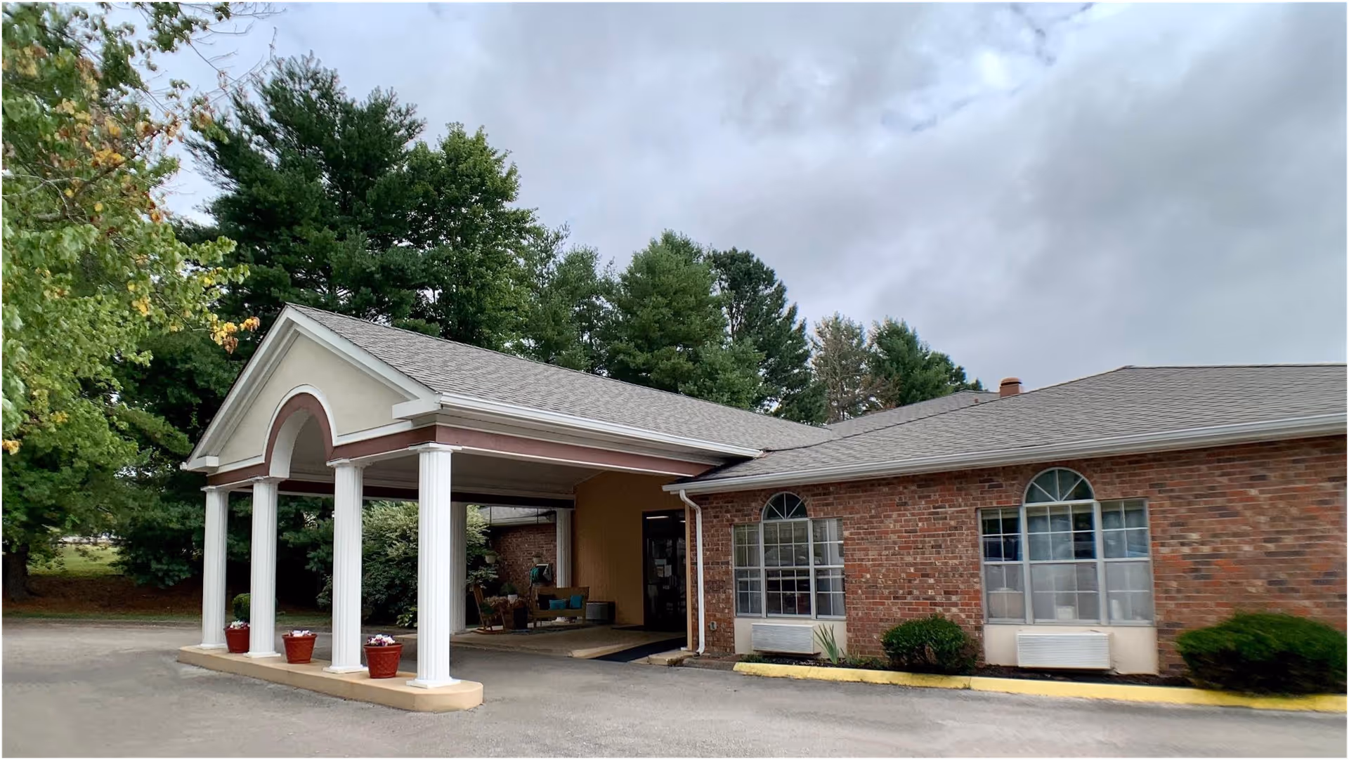 Front entrance of a single-story brick building with a covered portico supported by white columns, potted plants, and arched windows.