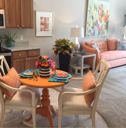 A small round orange dining table set with colorful place settings sits in an open-plan living area with a pink sofa and large floral artwork on the wall.
