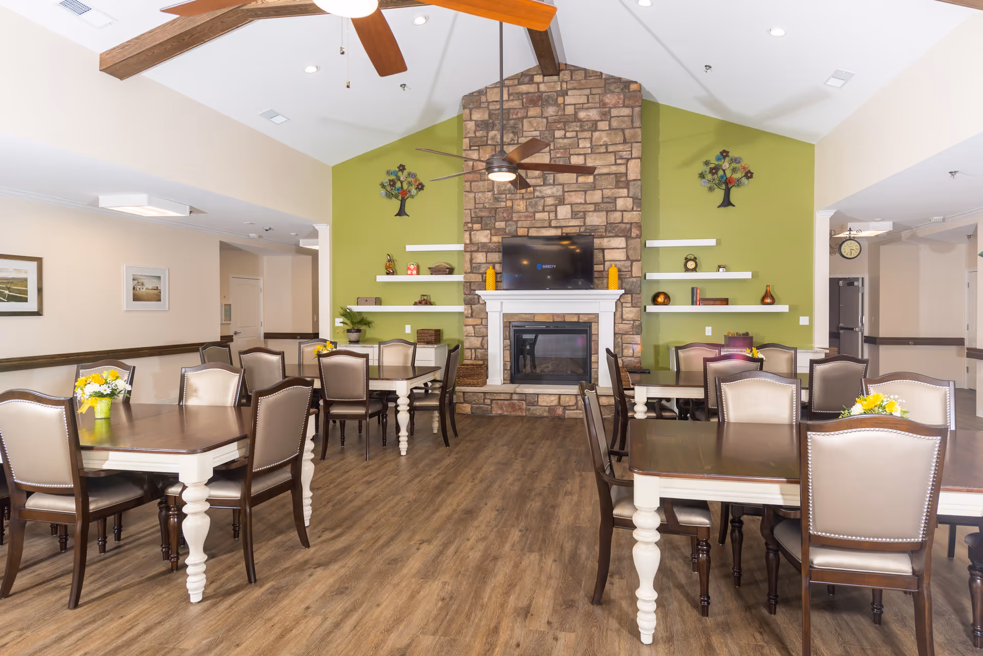 Communal dining room with multiple tables and chairs facing a stone fireplace and TV on a green accent wall.
