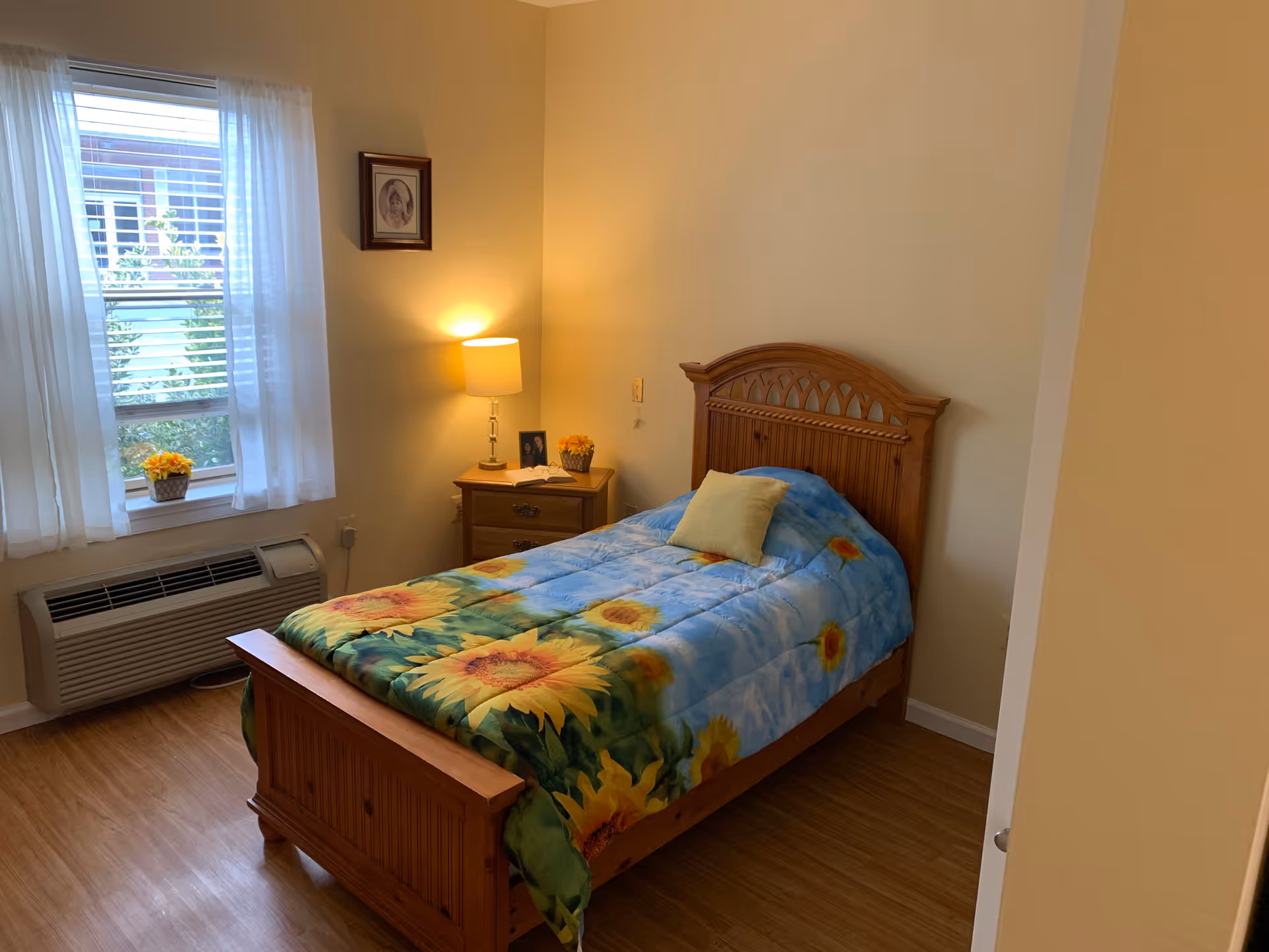 A cozy bedroom with a single wooden bed featuring a sunflower-themed comforter and a yellow pillow. Next to the bed is a wooden nightstand with a lit lamp, a small framed photo, and a flower pot. A window with white curtains allows natural light into the room, and below the window is an air conditioning unit. The floor is wooden, and the walls are painted a light beige color.