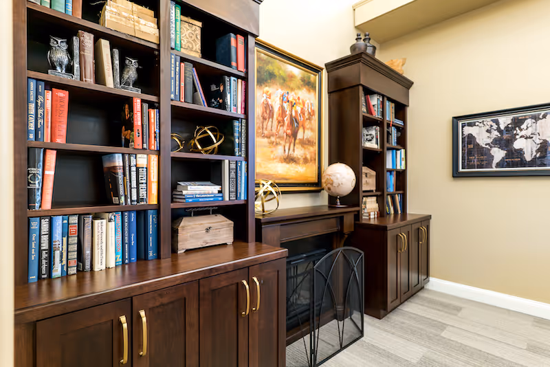 Well-lit interior room with dark wood bookshelves filled with books and decor, a small fireplace, a globe, and framed artwork on the walls.