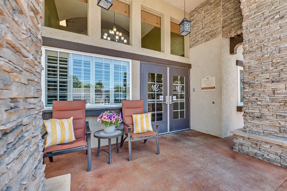Covered outdoor seating area at the entrance of Pacifica Senior Living South Coast featuring two cushioned chairs with striped pillows, a small round table with a flower pot, stone walls, and double glass doors with decorative logos.