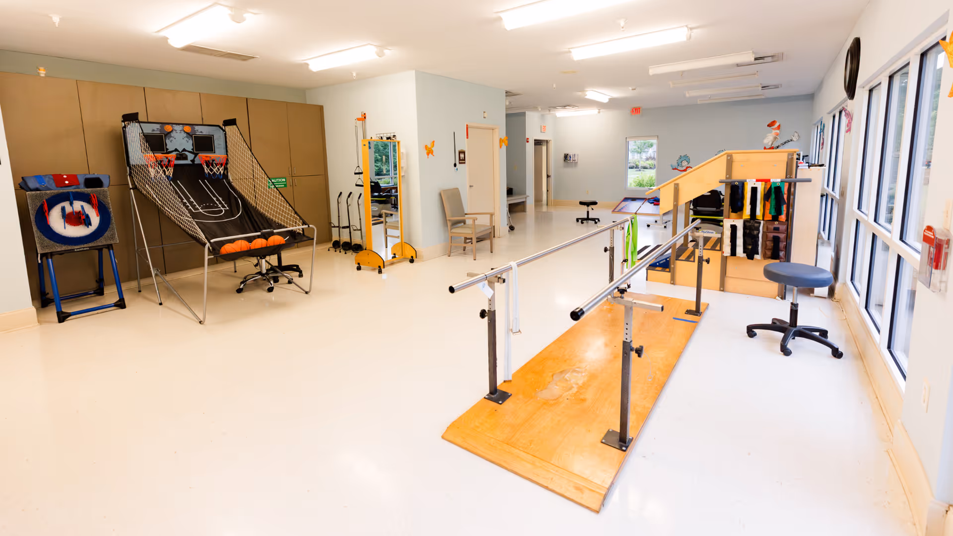 A spacious rehabilitation room with parallel bars on a wooden platform for walking exercises, a basketball arcade game, a cornhole game, chairs, and various physical therapy equipment. The room has large windows on one side allowing natural light to enter.