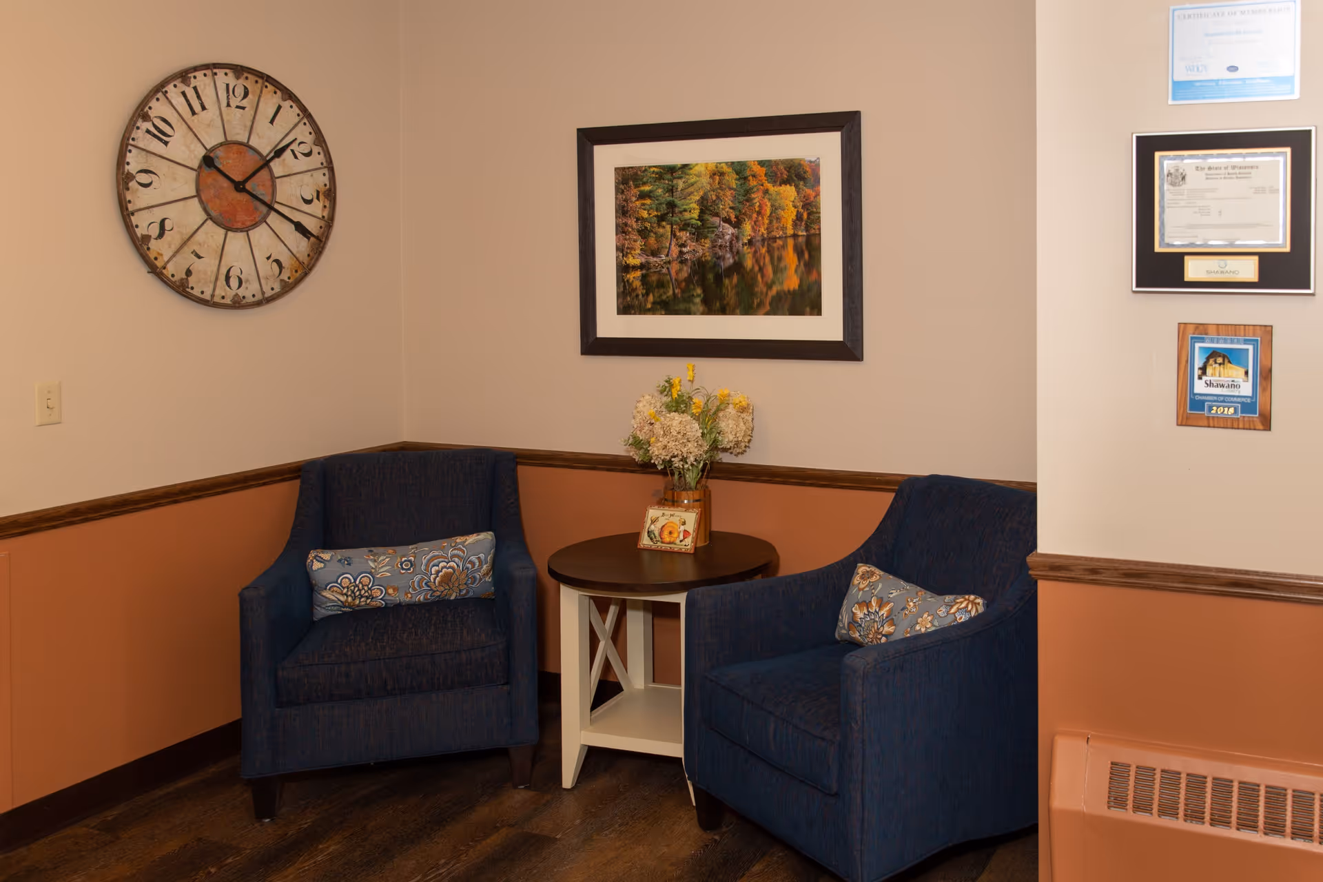 A cozy corner seating area with two blue upholstered armchairs, each with a decorative floral pillow. Between the chairs is a small round wooden table with a vase of flowers and a small framed picture. On the wall above the table is a framed photograph of a forest scene with autumn foliage reflected in water. To the left, a large vintage-style wall clock is mounted. On the right wall, several framed certificates and plaques are displayed. The walls are painted beige on the upper half and a warm brown on the lower half, separated by a wooden chair rail. The floor is dark wood.